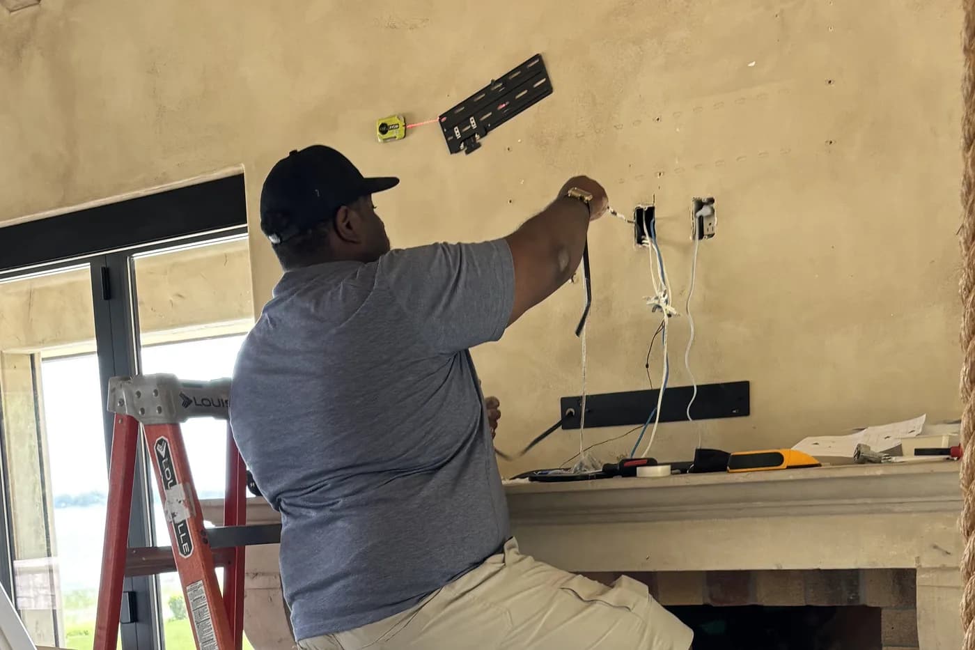 A technician on a ladder dressing cable behind a display wall-mount above a stone fireplace