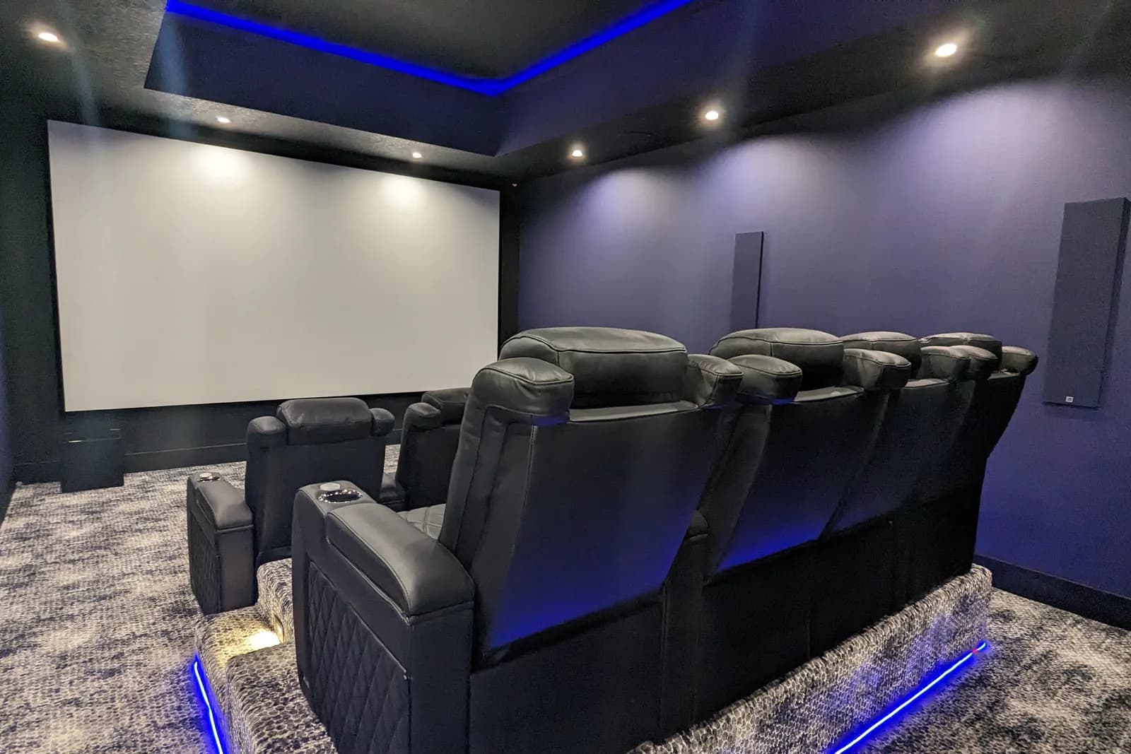 Cinema room with charcoal walls, blue LED cove lighting, black quilted recliners with lit bases and a wet bar at the rear